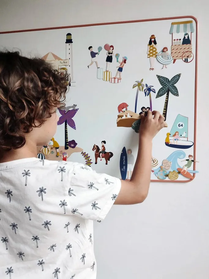 Un enfant de dos qui place des magnets illustrés de bord de mer sur un tableau magnétique accroché au mur