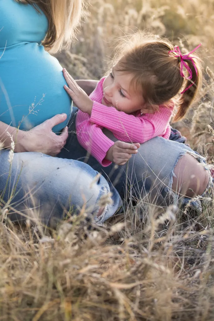 Une petite fille en pull rose qui pose les mains sur le ventre de sa mère enceinte, assises dans les herbes hautes