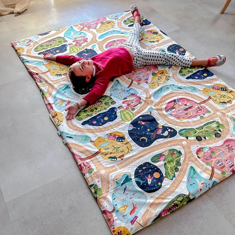 Un enfant allongé bras et jambes écartés sur un grand tapis d'éveil imprimé de routes et de maisons, posé sur un sol gris