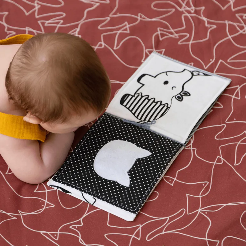 Un bébé à plat ventre sur un tapis d'éveil bordeaux imprimé de silhouettes blanches, qui regarde un livre en tissu noir et blanc