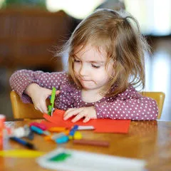 Une petite fille qui découpe du papier de couleur avec des ciseaux, à une table couverte de feutres
