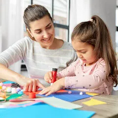 Une maman et sa fille penchées sur une table de bricolage, qui collent des papiers de couleur