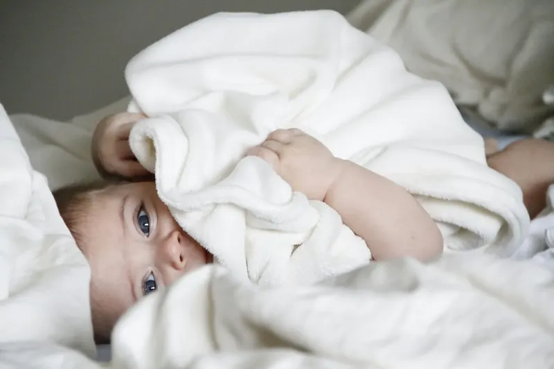 Un bébé allongé sur le côté, à moitié caché sous une couverture blanche, qui regarde l'objectif