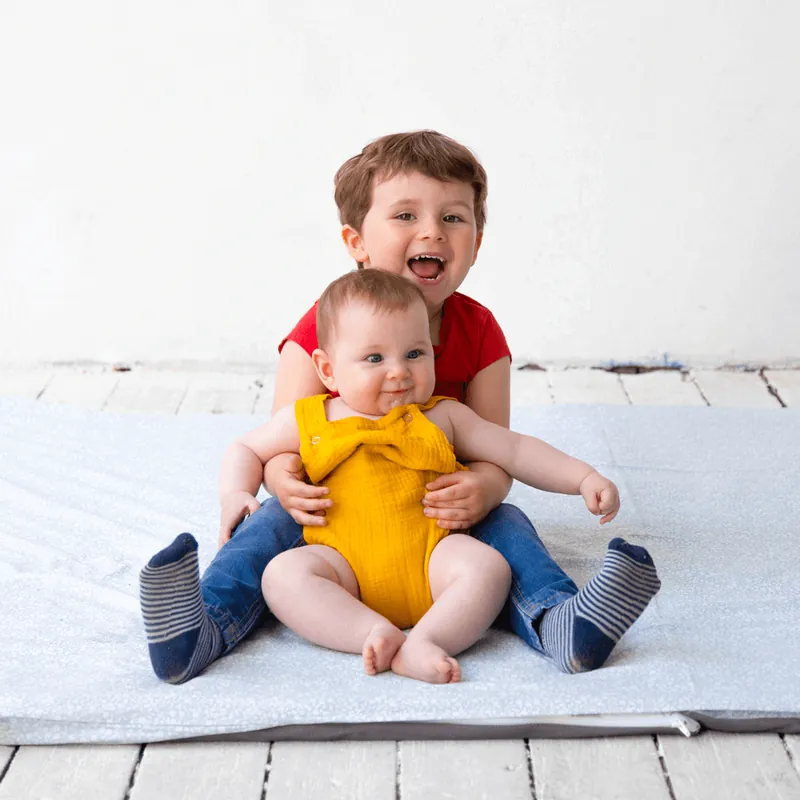 Un petit garçon qui rit, assis sur un tapis d'éveil gris clair, un bébé en barboteuse jaune sur les genoux