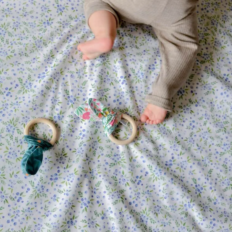 Deux anneaux de dentition en bois noués de tissu, posés sur un tapis à petites fleurs bleues près des jambes d'un bébé