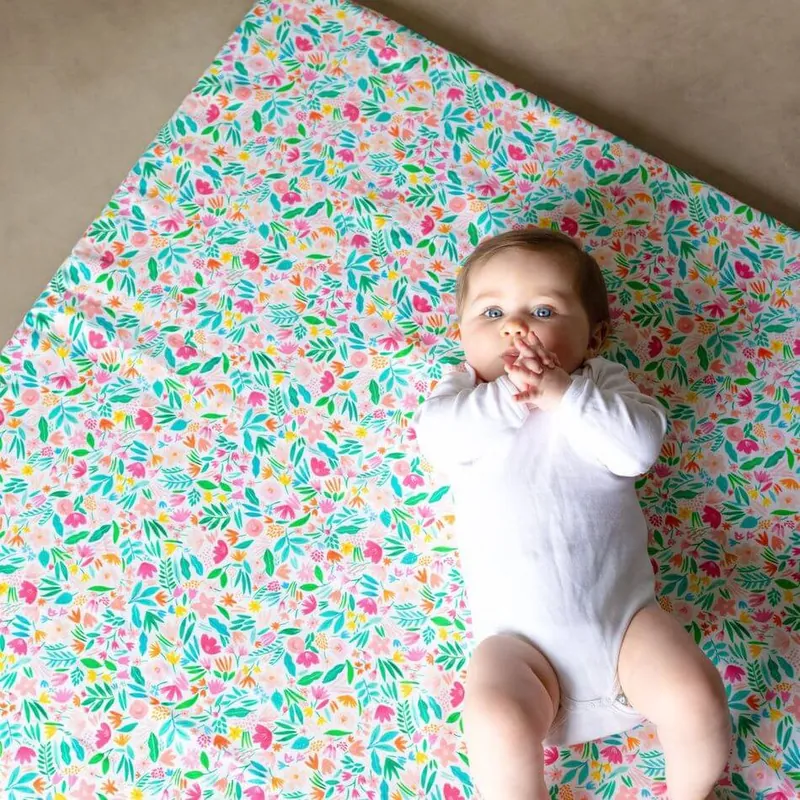Un bébé en body blanc allongé sur le dos sur un tapis d'éveil imprimé de fleurs roses et vertes, les mains à la bouche