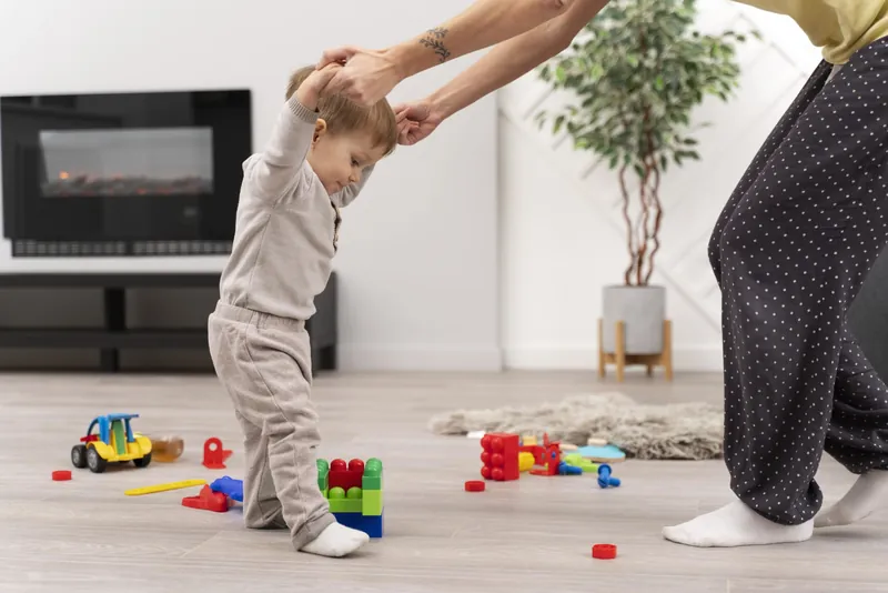 Un petit enfant qui fait ses premiers pas, tenu par les mains par un adulte, dans un salon aux jouets éparpillés