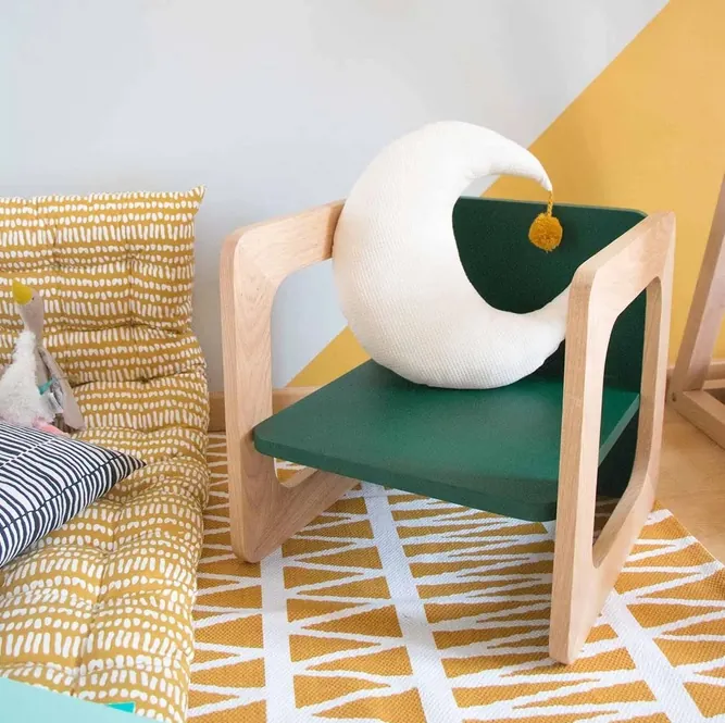 Un petit fauteuil d'enfant en bois à assise verte, un coussin en forme de lune posé dessus, sur un tapis jaune et blanc