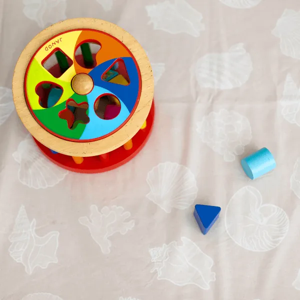 Une boîte à formes en bois aux couleurs vives et deux formes bleues posées sur un tapis rose pâle imprimé de coquillages