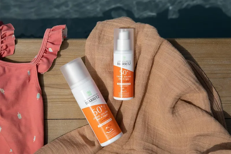Deux flacons de crème solaire bio indice 50+ des Laboratoires de Biarritz posés sur un lange, à côté d'un maillot de bain rose