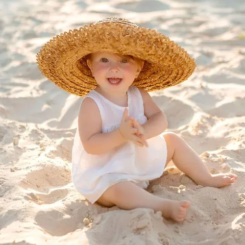 Une petite fille en robe blanche et grand chapeau de paille, assise dans le sable, qui sourit