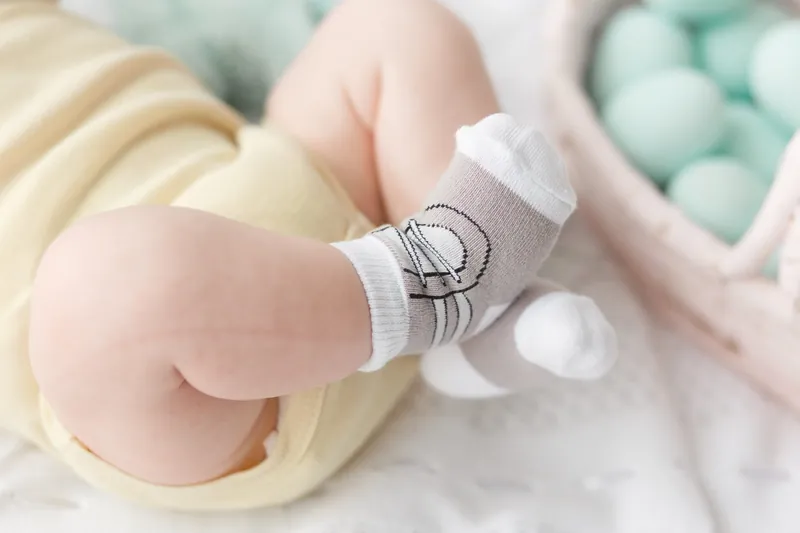 Les jambes d'un bébé en body jaune pâle et chaussettes grises, allongé sur une couverture blanche