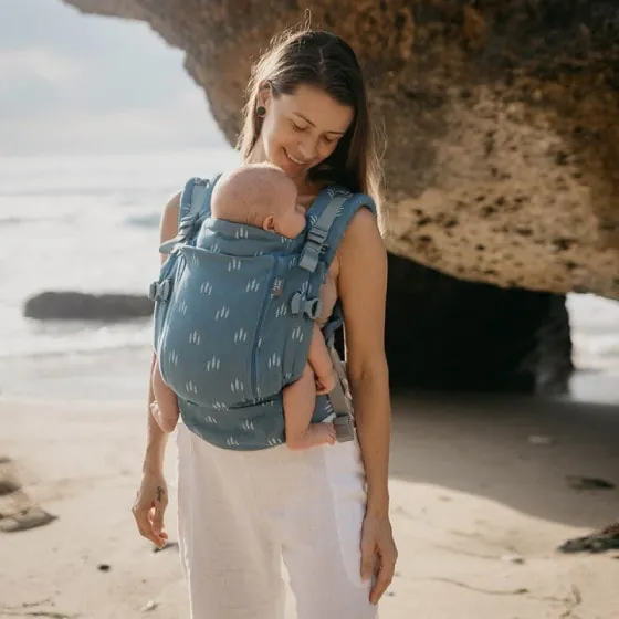 Une femme sur une plage qui porte son bébé contre elle dans un porte-bébé bleu à motifs