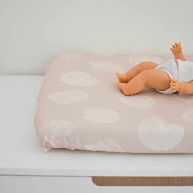 Un poupon allongé sur un matelas à langer rose pâle imprimé de coquillages blancs, posé sur un meuble blanc