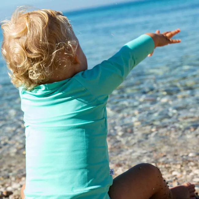Un enfant aux cheveux bouclés, en tee-shirt anti-UV turquoise à manches longues, assis au bord de l'eau, le bras tendu vers la mer