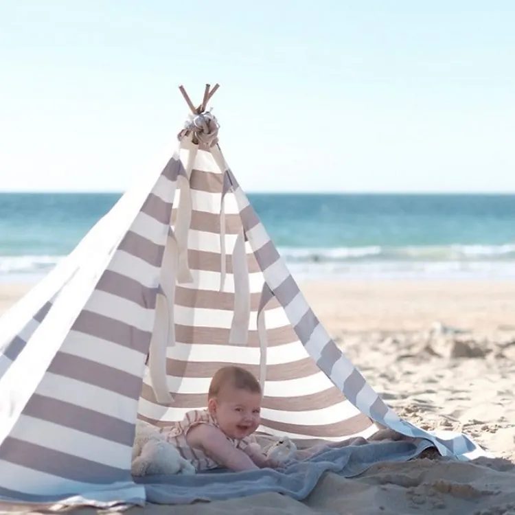 Un bébé allongé à plat ventre sous une tente de plage à rayures grises et blanches, face à la mer