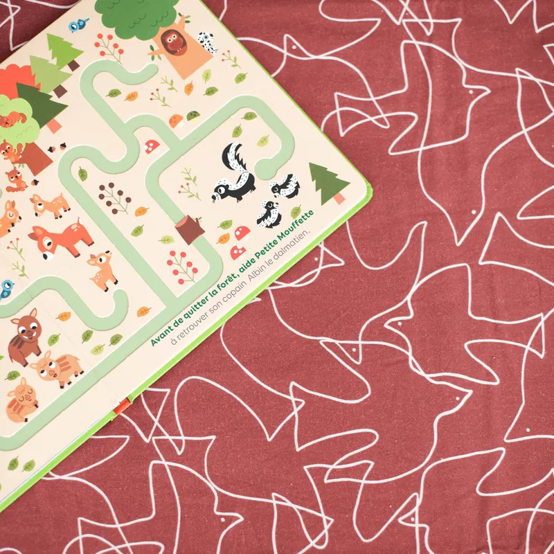 Un livre-jeu cartonné illustré d'animaux de la forêt, posé sur un tapis d'éveil bordeaux imprimé de silhouettes blanches