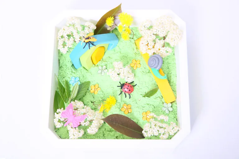 Un bac sensoriel blanc garni de sable vert, de fleurs, de feuilles et de petits animaux