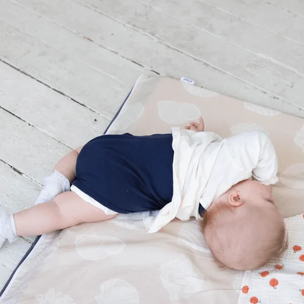 Un bébé en body bleu marine allongé sur le ventre, la joue contre un tapis d'éveil rose pâle imprimé de coquillages