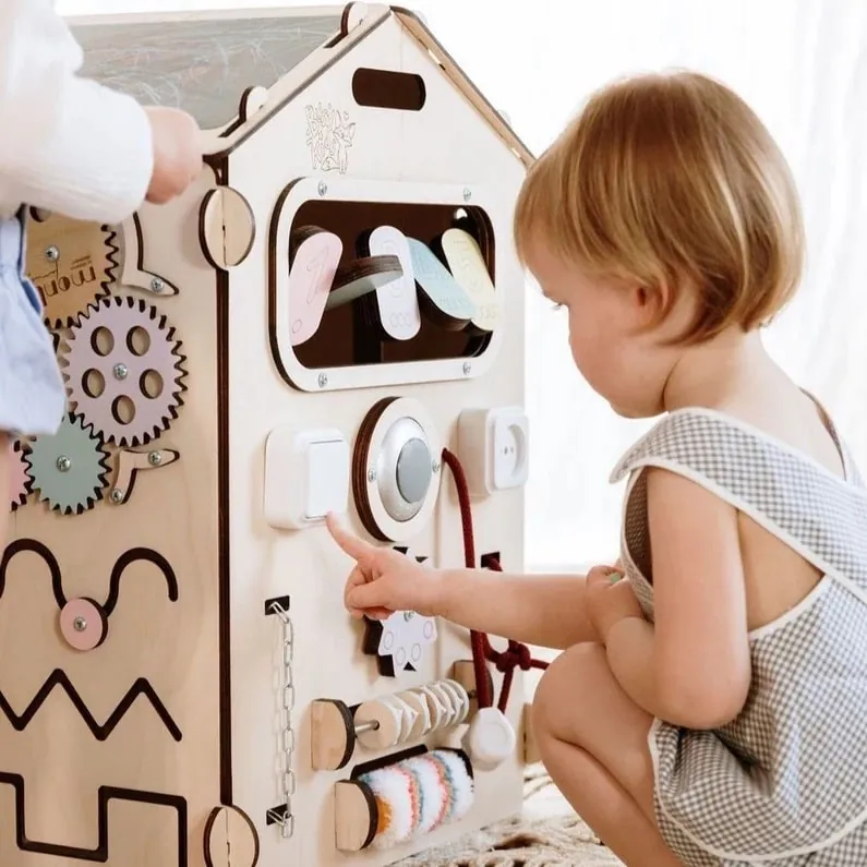 Une petite fille qui manipule les boutons et les engrenages d'un tableau d'activités en bois en forme de maison
