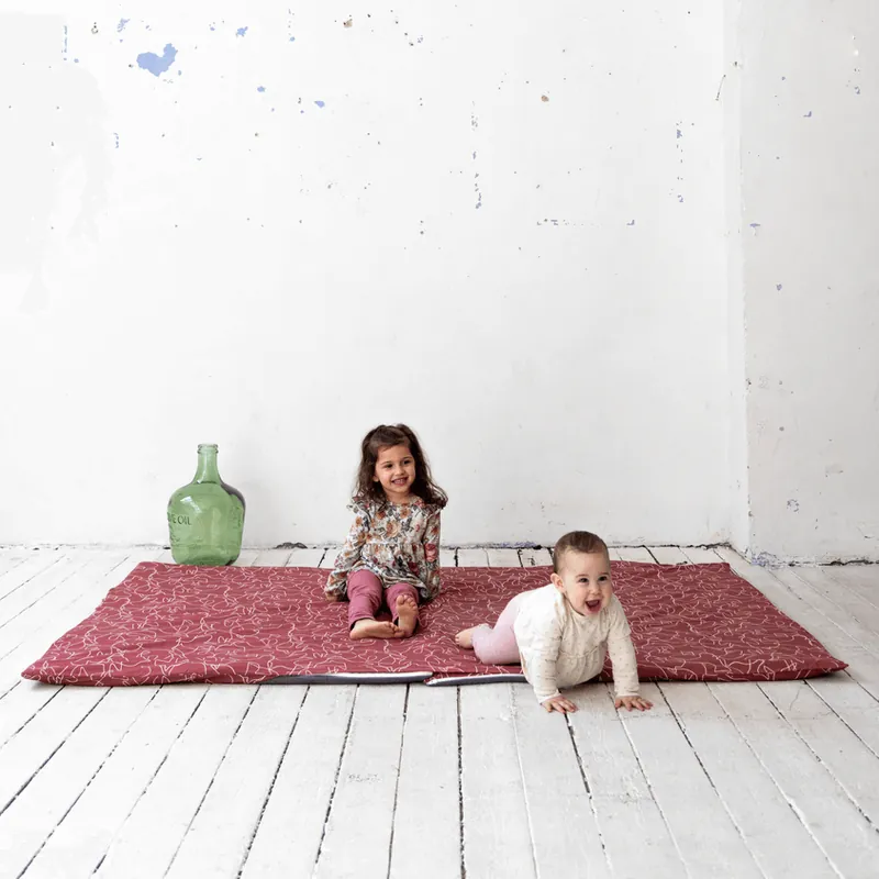 Une petite fille assise et un bébé à quatre pattes sur un tapis d'éveil bordeaux, dans une pièce blanche au parquet blanc