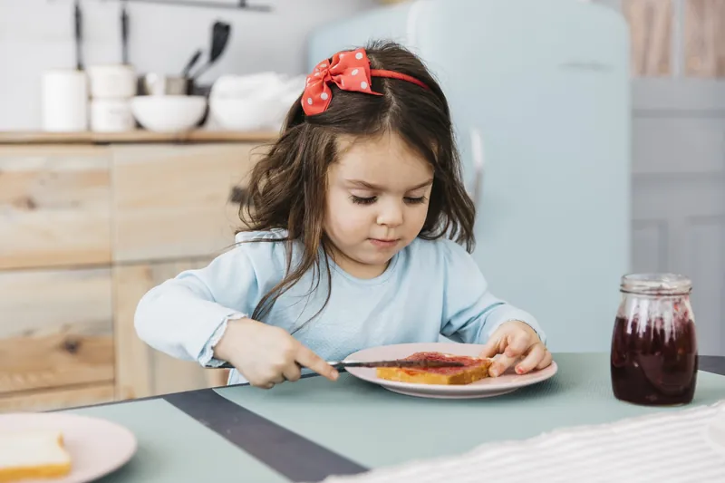 Une petite fille avec un nœud rouge dans les cheveux qui étale de la confiture sur une tartine