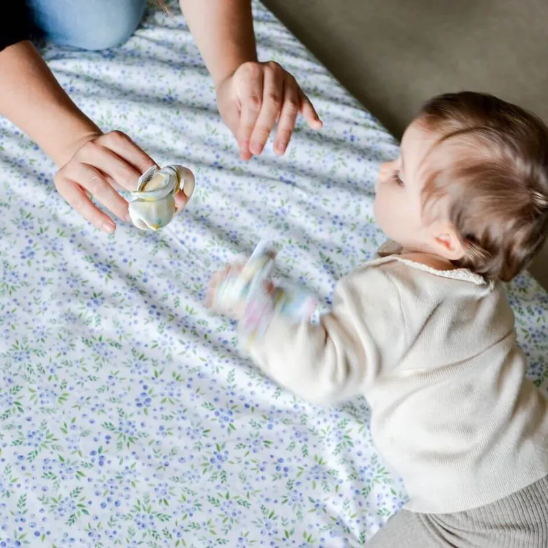 Un bébé sur un tapis d'éveil blanc à petites fleurs bleues, qui tend la main vers un anneau de dentition qu'un adulte lui présente