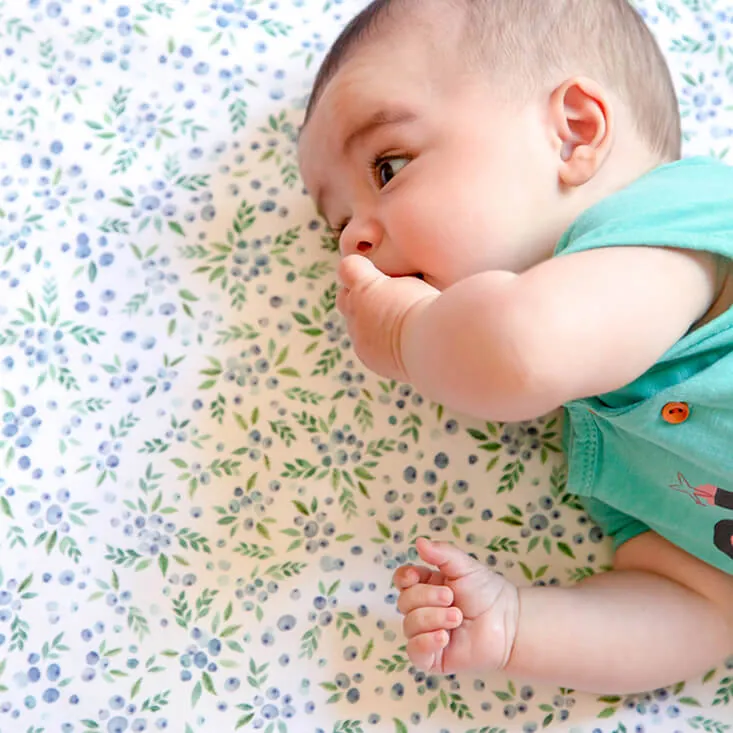 Un bébé allongé sur le côté qui suce son poing, sur un tapis d'éveil blanc imprimé de feuillages et de myrtilles