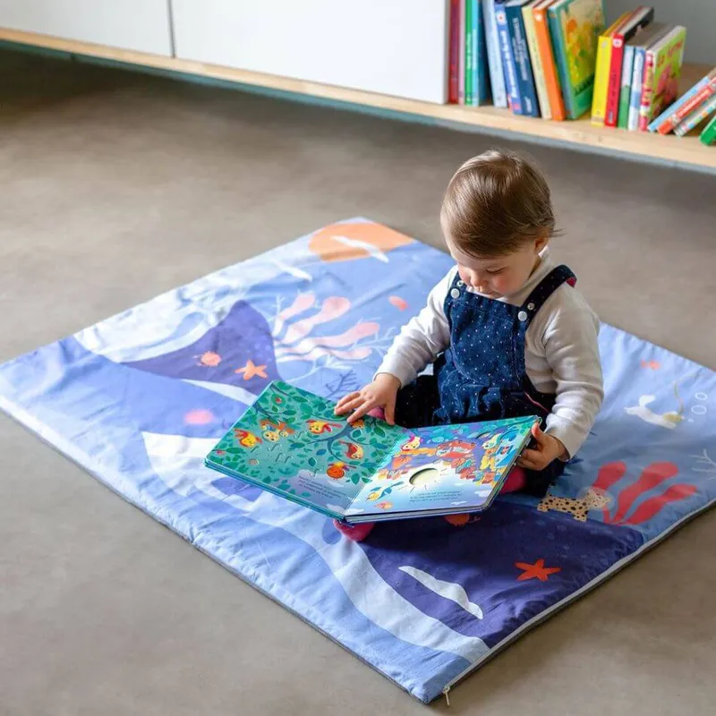 Un petit garçon en salopette assis sur un tapis d'éveil bleu imprimé de fonds marins, qui lit un livre illustré