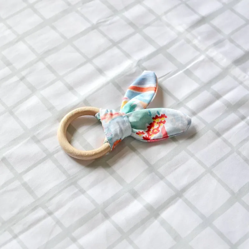 Un anneau de dentition en bois avec un noeud de tissu imprimé en forme d'oreilles de lapin, posé sur un drap blanc