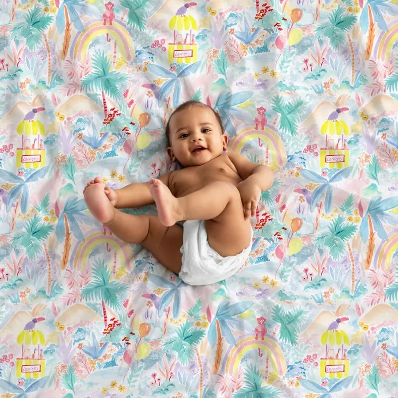 Un bébé souriant allongé sur le dos sur un tapis d'éveil imprimé de palmiers et d'arcs-en-ciel, qui attrape ses pieds