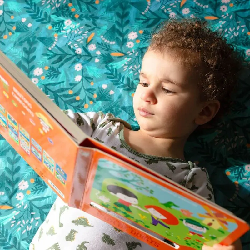 Un petit garçon allongé sur un tapis d'éveil imprimé de feuillages sur fond vert, qui lit un livre illustré