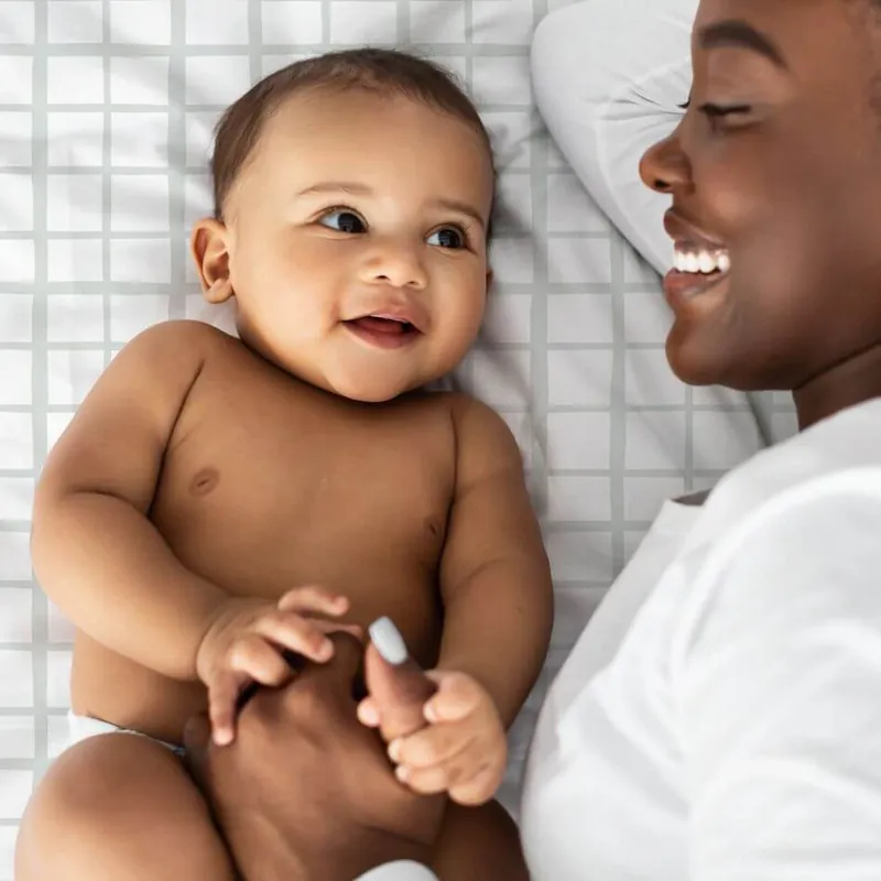 Un bébé souriant allongé sur un drap blanc, contre sa maman qui rit