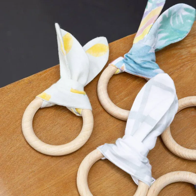 Des anneaux de dentition en bois avec des oreilles de lapin en tissu imprimé, posés sur une table en bois