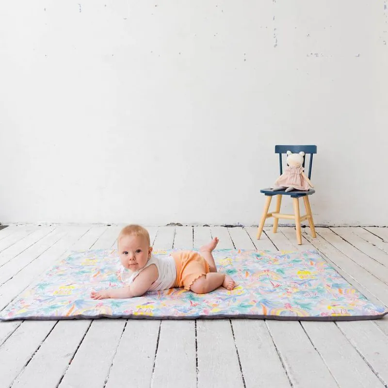 Un bébé allongé sur le ventre sur un tapis d'éveil aux motifs pastel, posé sur un parquet blanc, devant une petite chaise où est assise une peluche
