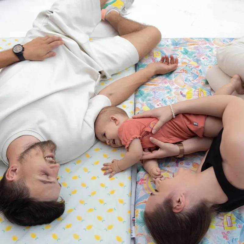 Un bébé allongé sur des tapis d'éveil imprimés, entre ses deux parents couchés au sol qui jouent avec lui