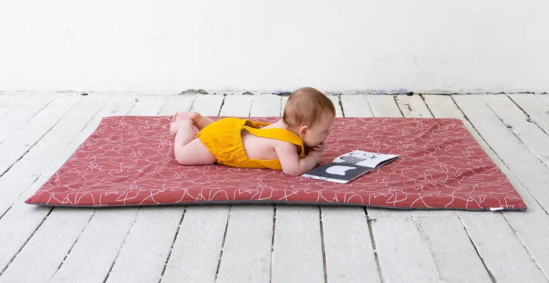 Un bébé allongé sur le ventre sur un tapis d'éveil rouge brique, qui regarde un livre en noir et blanc ouvert devant lui