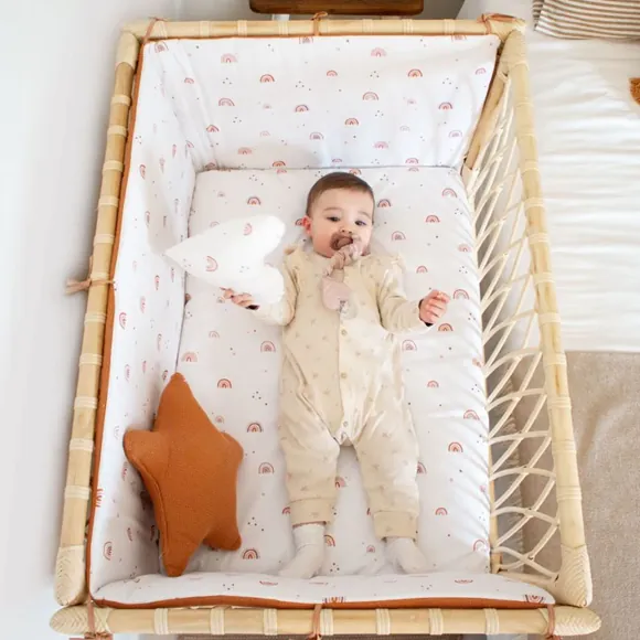 Un bébé allongé dans un lit cododo en rotin garni de draps imprimés d'arcs-en-ciel, à côté d'un coussin en forme d'étoile