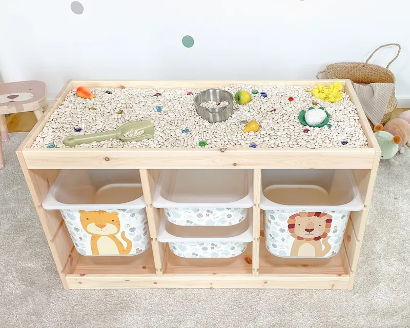 Une table d'activité sensorielle en bois remplie de graines et de petits objets, avec des bacs de rangement décorés d'animaux en dessous