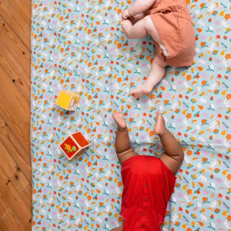 Vue de haut de deux bébés allongés tête-bêche sur un tapis d'éveil imprimé de fleurs sur fond bleu, près de deux cubes de jeu