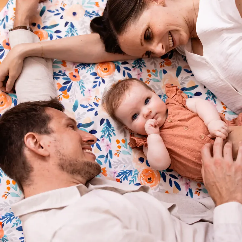 Un bébé allongé sur un tapis d'éveil imprimé de fleurs, entre son père et sa mère couchés près de lui qui lui sourient