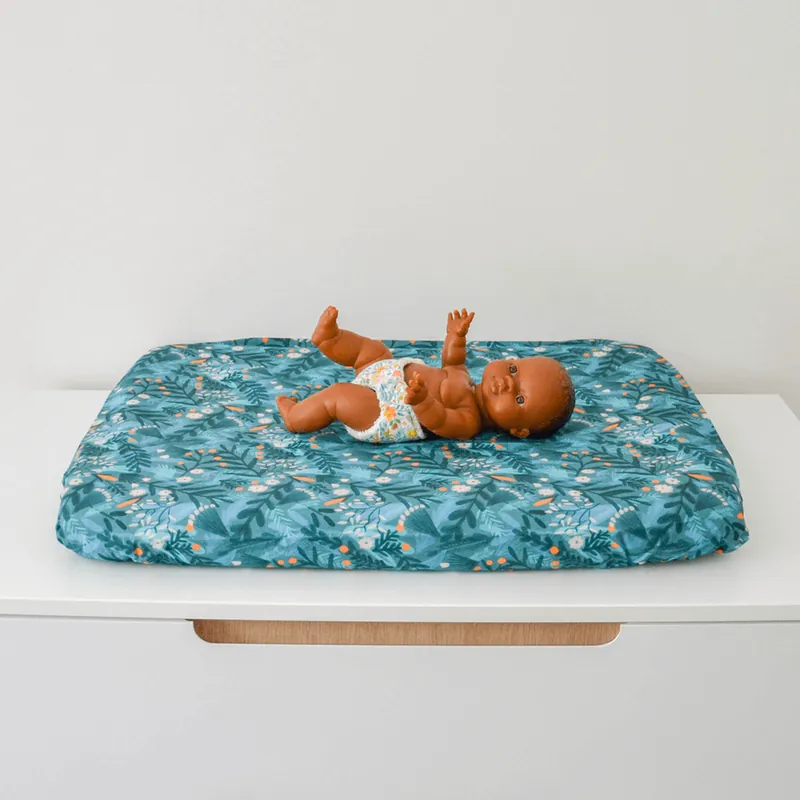 Un poupon posé sur un matelas à langer imprimé de feuillages sur fond bleu canard, sur une commode blanche