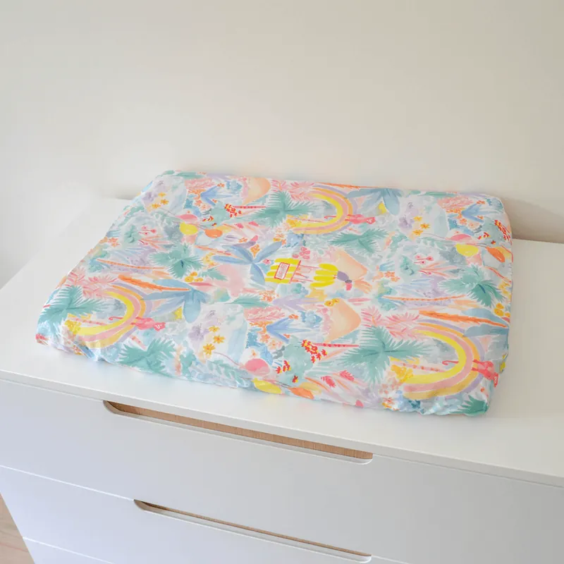 Un matelas à langer imprimé d'une jungle pastel aux arcs-en-ciel, posé sur une commode blanche