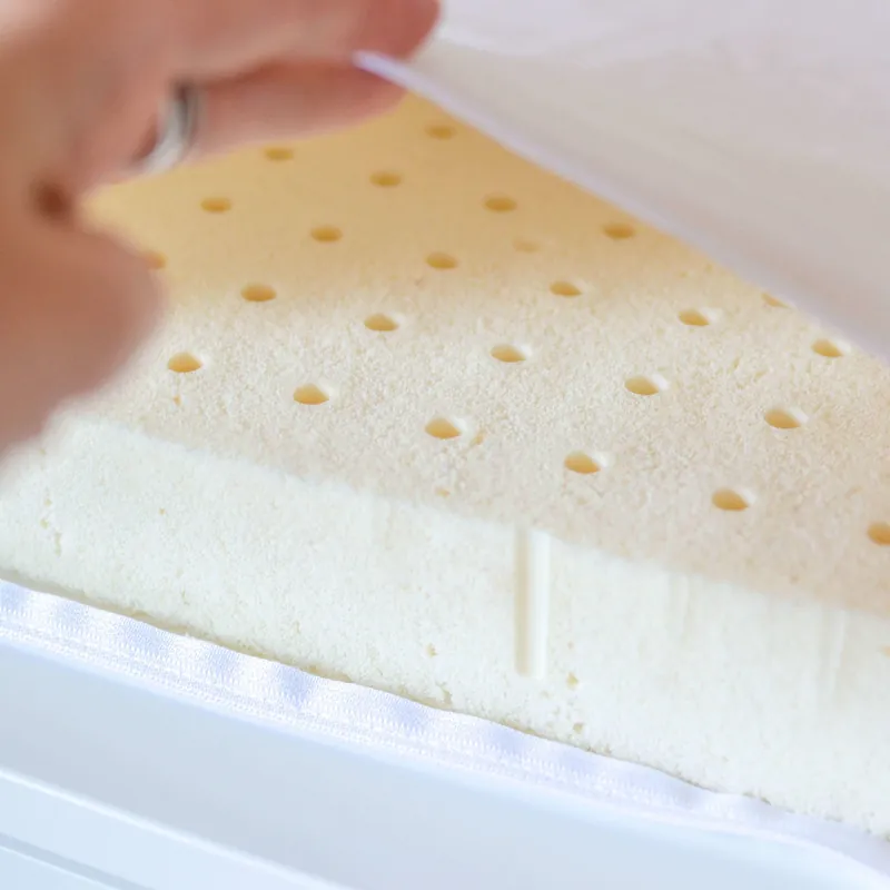Une main qui soulève l'alèse blanche d'un matelas à langer et montre la mousse en latex naturel perforée