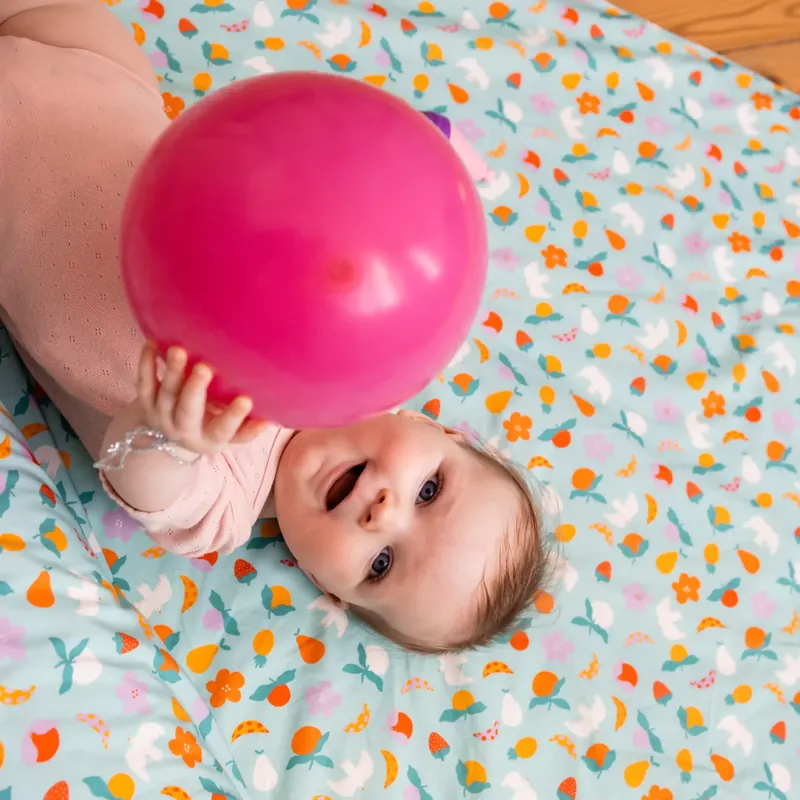 Un bébé allongé sur le dos qui tient un ballon rose à deux mains, sur un tapis d'éveil vert d'eau imprimé de petits fruits