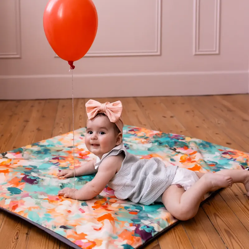 Un bébé avec un nœud rose dans les cheveux, allongé sur le ventre sur un tapis d'éveil aux couleurs d'aquarelle, sous un ballon rouge, dans une pièce rose