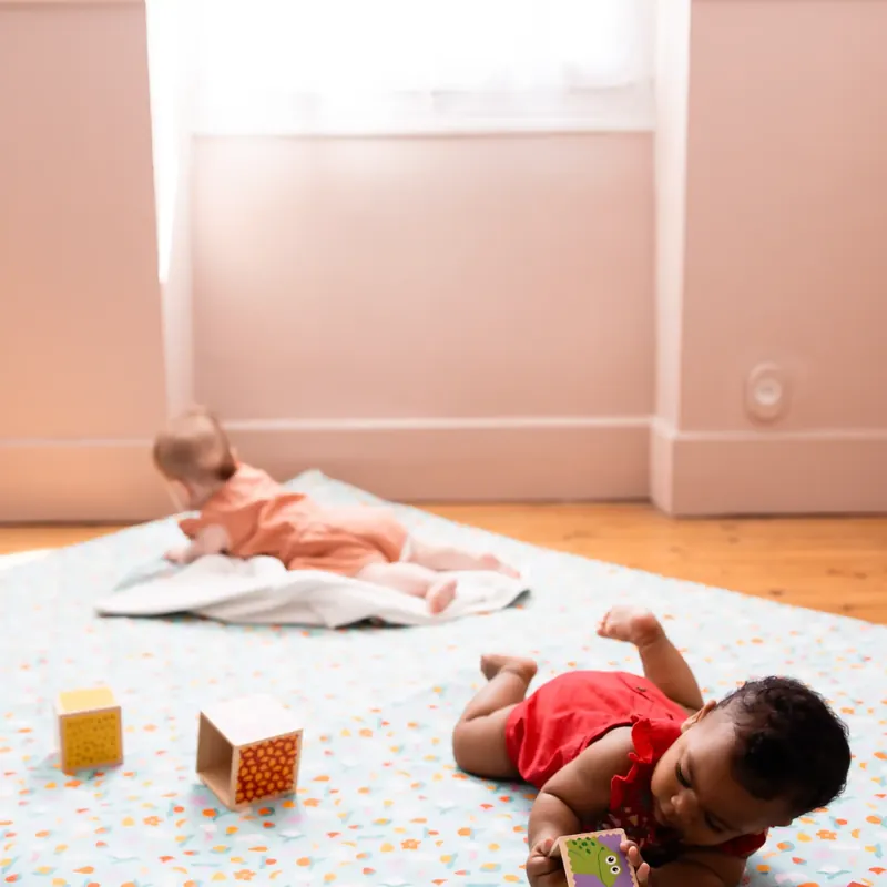 Deux bébés sur un grand tapis d'éveil blanc à petits motifs, l'un allongé au fond, l'autre qui joue avec des cubes et une boîte à formes en bois, dans une pièce rose