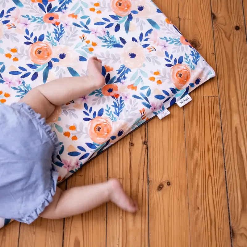 Les jambes d'un bébé en bloomer bleu sur le coin d'un tapis d'éveil blanc imprimé de grosses fleurs orange et bleues, posé sur un parquet
