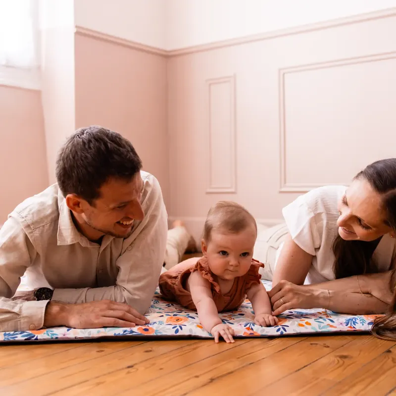 Un papa et une maman allongés sur le ventre de part et d'autre de leur bébé, sur un tapis d'éveil imprimé posé sur un parquet