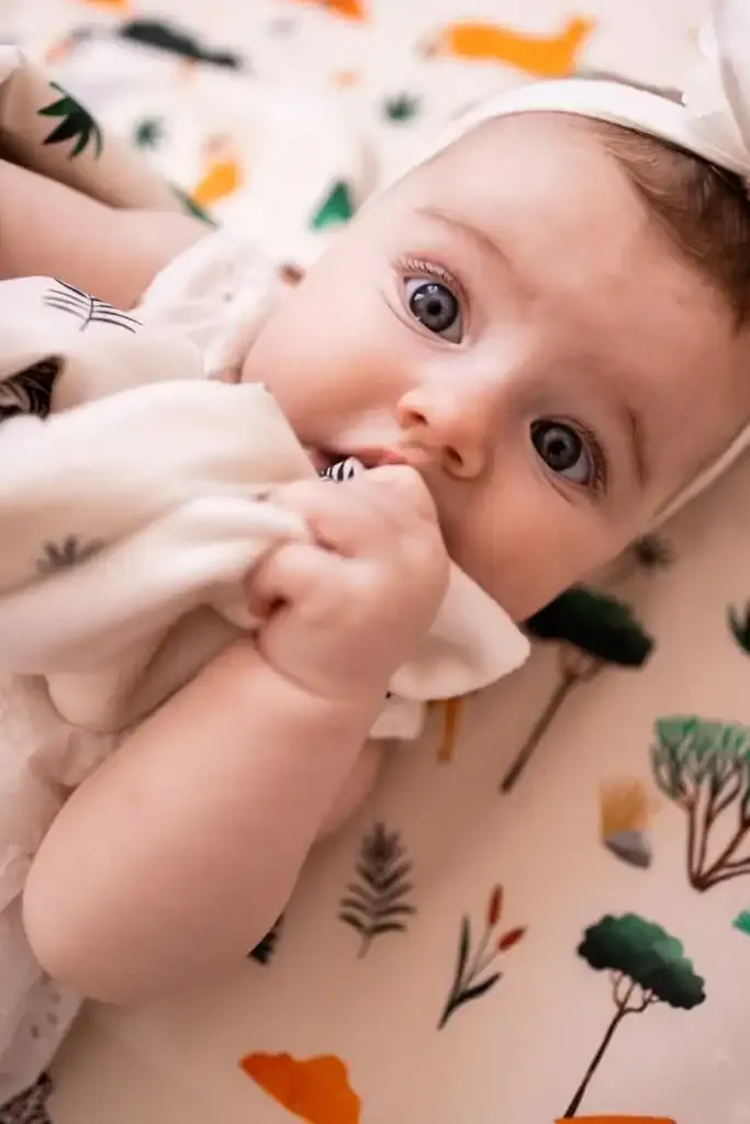 Un bébé aux grands yeux qui mordille un doudou blanc, allongé sur un tapis d'éveil blanc imprimé de petits arbres